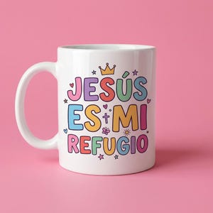 May include: White ceramic mug with the text "JES&Uacute;S ES MI REFUGIO" in colorful bubble letters. The design includes a crown, stars, hearts, and flowers. The mug has a curved handle and is set against a pink background.