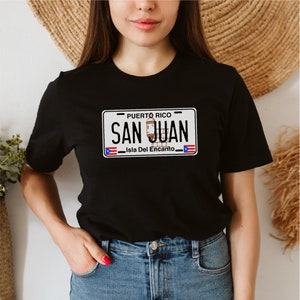 May include: A black t-shirt with a white graphic of a Puerto Rican license plate that says "PUERTO RICO SAN JUAN Isla Del Encanto".