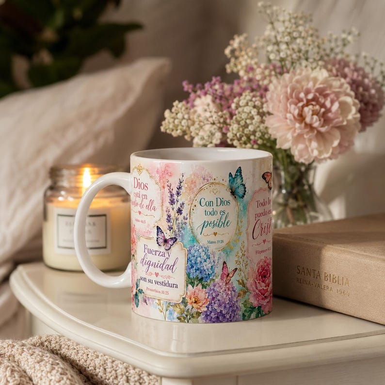 May include: White ceramic mug with floral and butterfly designs, and text in Spanish. The mug has a handle and is on a white surface next to a candle and a vase of flowers. A book is also visible.