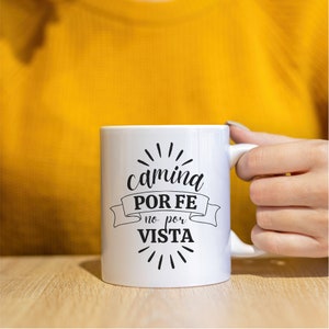 May include: White ceramic mug with black text that reads "camina POR FE no por VISTA" with a black ribbon design and black starburst design.