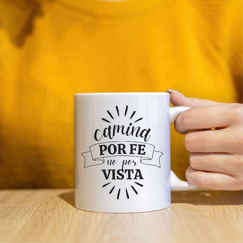 May include: White ceramic mug with black text that reads "camina POR FE no por VISTA" with a black ribbon design and black starburst design.