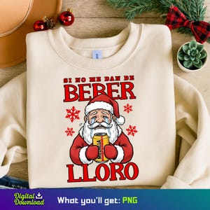 May include: A cream-coloured sweatshirt featuring a crying Santa Claus design. The Santa is depicted in a red suit and hat, clutching a beer can. The text on the shirt reads "SI NO ME DAN DE BEBER LORO" in red, with snowflake accents.