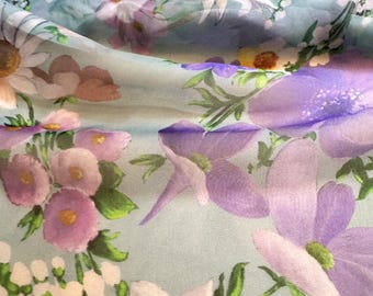 Op de afbeelding: Close-up van een stof met een bloemenpatroon. De stof heeft een lichtblauwe achtergrond met een aquarel-achtig ontwerp van paarse, witte en roze bloemen. De bloemen zijn van verschillende groottes en vormen, met groene bladeren.