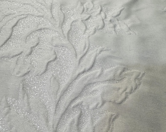 May include: Close-up of a white fabric with an embossed floral pattern. The design features detailed flowers and leaves with a subtle, shimmering texture. The fabric appears soft and luxurious, suitable for home decor.