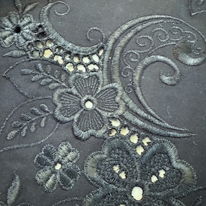 May include: Close-up of black fabric with intricate floral embroidery. The design features detailed flowers, leaves, and swirling patterns, creating a textured and elegant appearance. The embroidery is in a darker shade of black, providing a subtle contrast.