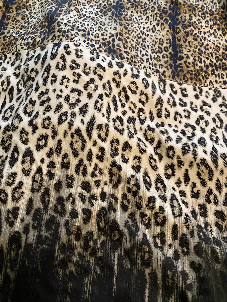 May include: A close-up of a fabric with a leopard print pattern. The fabric is brown, black, and white. The print is a classic leopard print with black spots on a brown background.