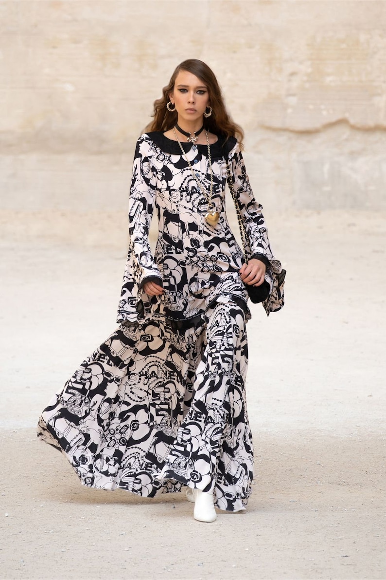 Peut inclure: Une femme portant une robe &agrave; motifs noir et blanc &agrave; manches longues et une jupe ample. Elle porte un collier en or avec un pendentif en forme de c&oelig;ur et des bottes &agrave; talons blanches.