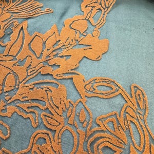 May include: Close-up of a textile featuring an intricate floral design in a warm orange hue. The design is overlaid on a teal fabric, creating a striking contrast. The pattern includes detailed cut-out shapes and textured elements.