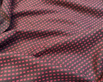 May include: Close-up of a dark fabric with a repeating pattern of small, bright red rectangles. The rectangles are arranged in a grid-like formation, creating a textured appearance. The fabric is slightly wrinkled, adding depth to the design.