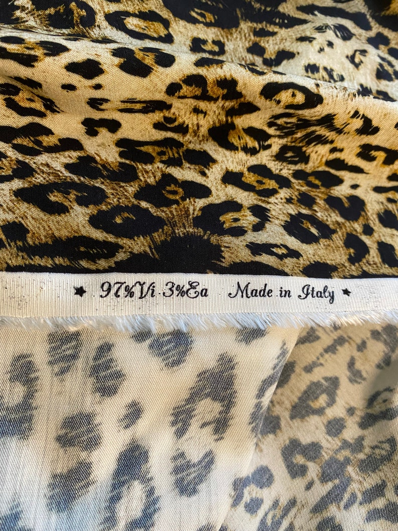 May include: Close-up of a fabric swatch with a leopard print pattern. The fabric is beige with black spots. The label reads "97% Vi - 3% Ea Made in Italy".