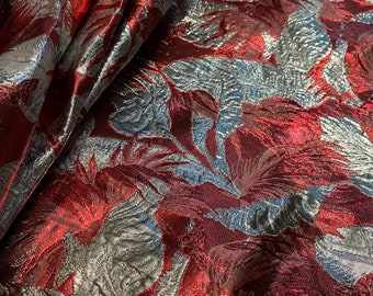 May include: Close-up of a luxurious fabric with a floral pattern. The fabric has a metallic sheen with deep red and silver colours. The design is a repeating pattern of stylized leaves and flowers.