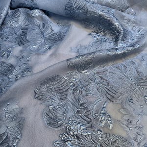 Könnte beinhalten: Ein hellblauer, durchsichtiger Stoff mit einem silbernen, metallischen Blumenmuster. Der Stoff ist strukturiert und hat einen dezenten Glanz.