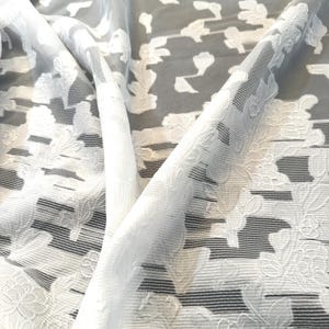 May include: White sheer fabric with a raised floral pattern. The fabric is textured and has a subtle sheen.