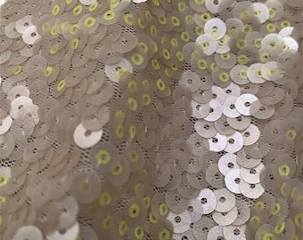 May include: A close-up of a fabric with a pattern of overlapping sequins. The sequins are a light beige color with a subtle shimmer and have a light yellow outline.