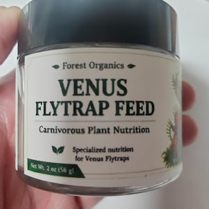 May include: A clear plastic jar with a black lid containing Forest Organics Venus Flytrap Feed. The label features green text reading Venus Flytrap Feed, Carnivorous Plant Nutrition, and Specialized nutrition for Venus Flytraps, with a net weight of 56 g.