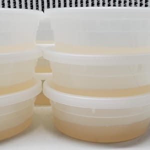May include: A set of six round, semi-transparent plastic food storage containers stacked in two columns of three. Each container features a matching white lid and is filled with a pale yellow liquid substance, showing a clear view of the contents.