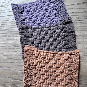 May include: Three woven coasters with fringed edges. The top coaster is purple, the middle coaster is gray, and the bottom coaster is brown.
