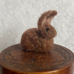 May include: A small, brown, handmade bunny figurine with a textured, woolly appearance. The bunny sits atop a wooden surface. The bunny has a curved ear and a small, dark eye. The figurine is a charming, handcrafted item.