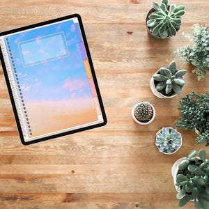 Pu&ograve; includere: Un tablet digitale che mostra un quaderno a spirale con una copertina a tema cielo caratterizzata da un gradiente blu e arancione. Il quaderno include un'etichetta rettangolare e schede laterali colorate su una superficie in legno.