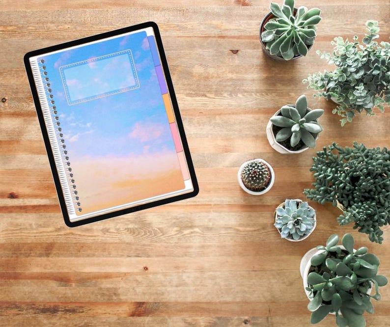 Pu&ograve; includere: Un tablet digitale che mostra un quaderno a spirale con una copertina a tema cielo caratterizzata da un gradiente blu e arancione. Il quaderno include un'etichetta rettangolare e schede laterali colorate su una superficie in legno.