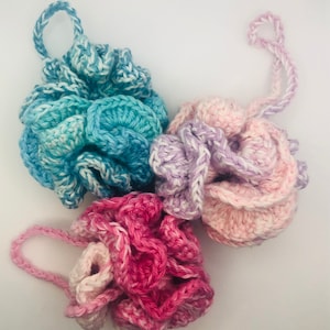 May include: Three handmade crochet bath poufs in shades of blue, pink, and purple. Each pouf has a loop for hanging. The crochet work is detailed, creating a textured, floral-like appearance. The poufs are arranged on a white background.