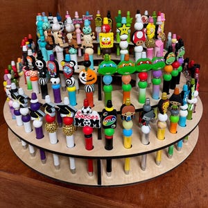 Könnte beinhalten: Ein mehrstöckiger Displayständer aus Holz, gefüllt mit bunten Stiften. Die Stifte haben dekorative Aufsätze mit verschiedenen Figuren, Formen und Designs. Der Ständer ist aus hellfarbenem Holz gefertigt und mit Stiften in verschiedenen Farben gefüllt.