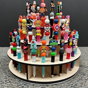 May include: A two-tiered wooden display stand filled with colorful pens. The pens have various decorative toppers, including cartoon characters, beads, and other novelty designs. The pens are arranged in a circular pattern.