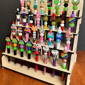 May include: A wooden display stand filled with colorful beaded pens. The pens feature various designs, including flowers, cartoon characters, and hearts. The stand has multiple tiers, showcasing the pens in an organized manner. The pens are in a variety of colors.