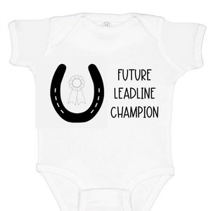 May include: White baby onesie with a black horseshoe and a ribbon inside. The text "FUTURE LEADLINE CHAMPION" is printed below the horseshoe.
