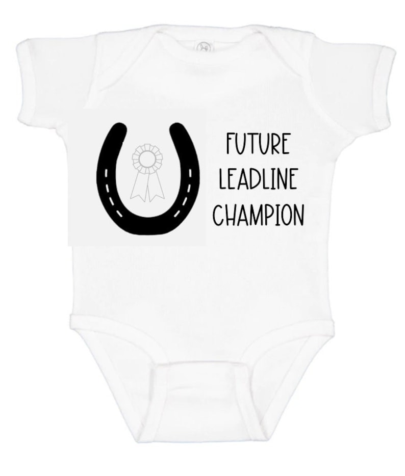 May include: White baby onesie with a black horseshoe and a ribbon inside. The text "FUTURE LEADLINE CHAMPION" is printed below the horseshoe.