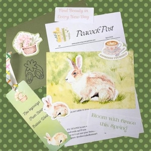 Puede incluir: Una colección de artículos de papelería con temática primaveral, que incluye una pintura de acuarela de un conejo, pegatinas y un marcapáginas. Los artículos presentan colores pastel y diseños florales, con el texto "Find Beauty in Every New Day."