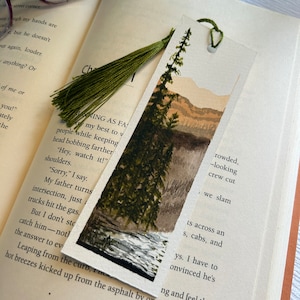 May include: A watercolor bookmark featuring a green forest scene with a river and mountains in the background. The bookmark has a green tassel attached to the top.