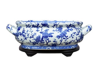 Könnte beinhalten: Ein weißer Keramik-Pflanztopf mit blauem Blumenmuster und einem schwarzen Holzsockel. Der Topf ist oval geformt und hat zwei Griffe an den Seiten.