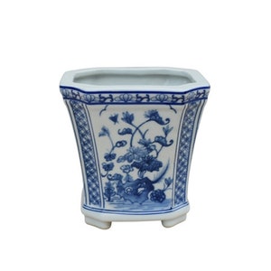 May include: A white ceramic planter with a blue and white floral design. The planter has a square shape with rounded corners and four small feet.