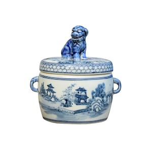 May include: A blue and white porcelain jar with a lid featuring a blue lion finial. The jar is decorated with a blue and white landscape scene.