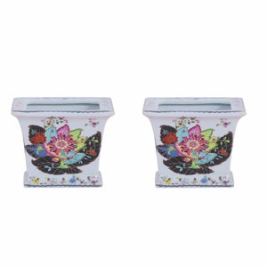 May include: Two white ceramic square planters with a floral design. The planters feature a large pink flower with green and blue leaves, surrounded by smaller flowers and leaves. The planters have a blue and gold trim.