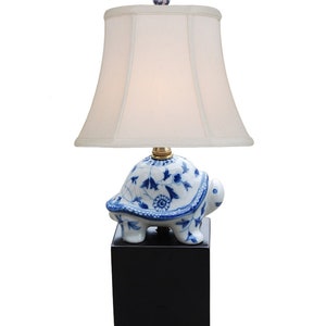 May include: A blue and white porcelain turtle lamp base with a white lampshade. The lamp base is on a black square base with rounded feet.