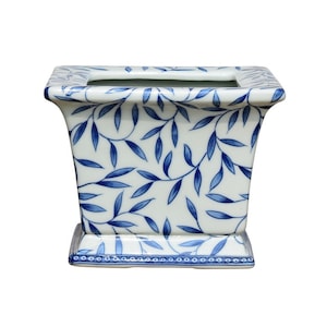 May include: A white ceramic planter with a blue and white leaf pattern. The planter has a square base and a rectangular body.
