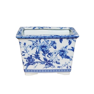 May include: A white ceramic planter with a blue floral pattern. The planter has a rectangular shape with rounded corners and four small feet.