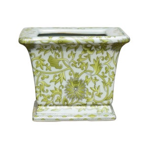 May include: A white ceramic planter with a green floral design. The planter has a square base and a rectangular body.