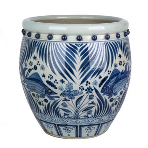 May include: A large, white ceramic planter with a blue and white design. The planter features a repeating pattern of stylized leaves and fish. The rim of the planter is decorated with a row of blue beads.