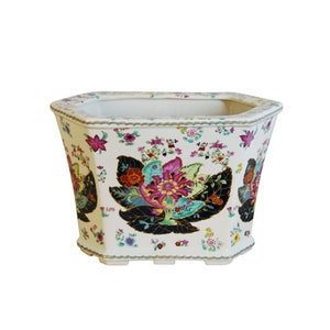 May include: White ceramic planter with a hexagonal shape and a floral design. The planter features a large central flower with pink, red, and green petals surrounded by smaller flowers and leaves in shades of blue, green, and black.