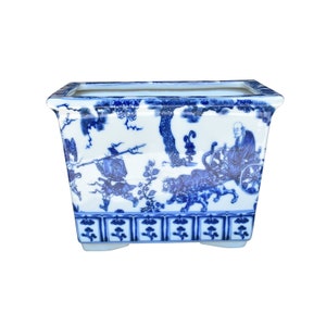 May include: A rectangular blue and white porcelain planter with a traditional Chinese design. The planter features a scene with a man riding in a cart pulled by a tiger, surrounded by trees and other figures.