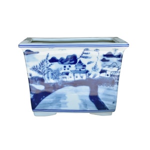 May include: A rectangular blue and white ceramic planter with a landscape scene painted on the front. The planter has a square base and a slightly flared top.