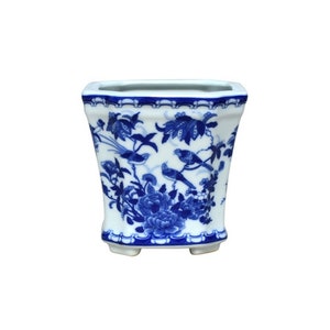 May include: A white ceramic planter with a blue and white floral design featuring birds. The planter has a square shape with rounded corners and a slightly flared lip.