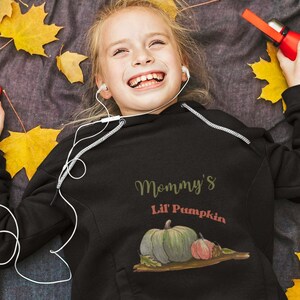 May include: A young person wearing a black hoodie with the text "Mommy's Lil Pumpkin" and a graphic of a green pumpkin with a smaller orange pumpkin beside it. The person is lying on a blanket with fall leaves.