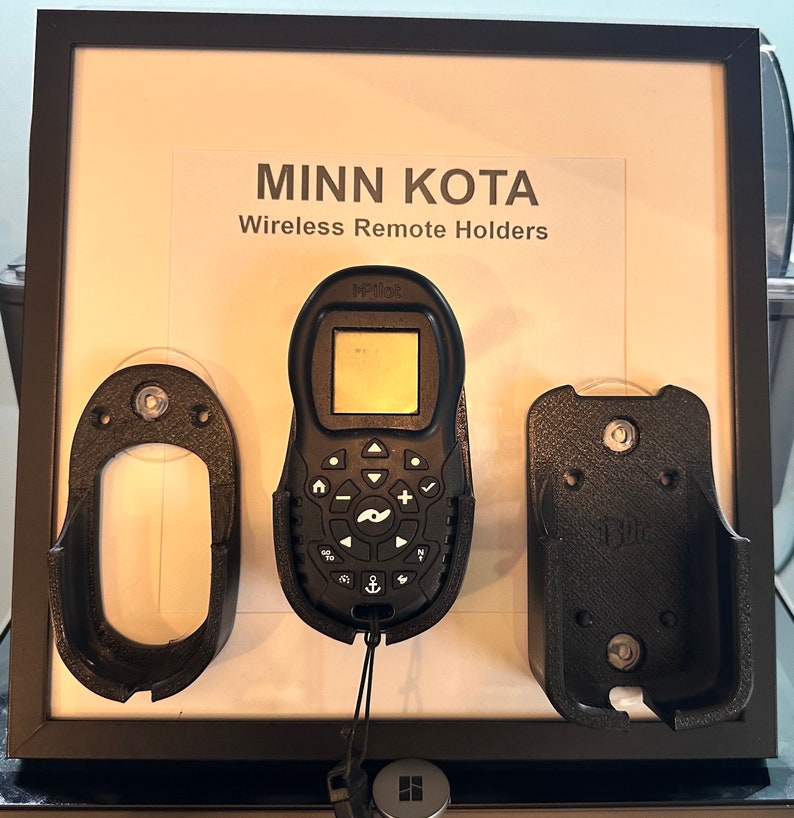 Puede incluir: Un tablero de exhibici&oacute;n blanco con el texto "MINN KOTA Wireless Remote Holders" y tres soportes de pl&aacute;stico negro. El soporte central tiene un control remoto negro con una pantalla y botones. El control remoto est&aacute; etiquetado como "i-Pilot".