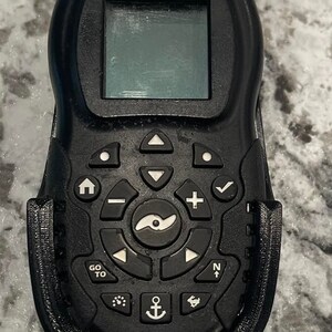 Puede incluir: Un mando a distancia iPilot negro con una peque&ntilde;a pantalla, botones y una palanca de mando. El mando est&aacute; sujeto a un soporte negro.