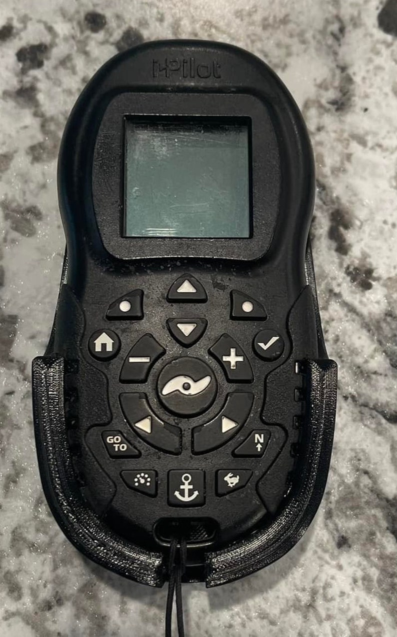 Puede incluir: Un mando a distancia iPilot negro con una peque&ntilde;a pantalla, botones y una palanca de mando. El mando est&aacute; sujeto a un soporte negro.