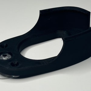 Puede incluir: Un soporte de pared de impresi&oacute;n 3D negro con una almohadilla adhesiva transparente en la parte inferior. El soporte tiene una forma curva y una abertura circular en el centro.